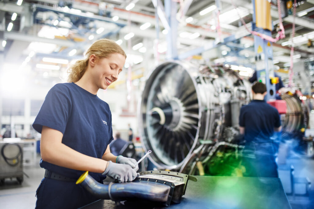 Dieses Kampagnenfoto der Lufthansa Technik zeigt eine Mechanikerin, die an einem Triebwerkteil arbeitet. Im Hintergrund zu sehen ist ein weiterer Mitarbeiter, der an einem Flugzeugtriebwerk arbeitet.