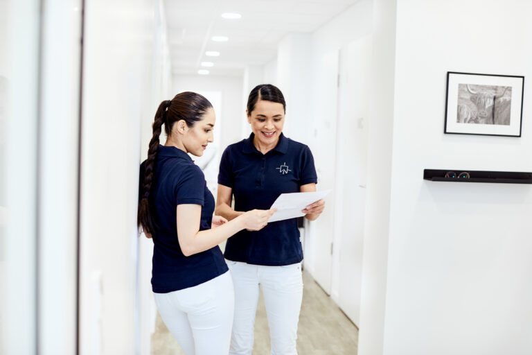 Zwei Sprechstundenhilfen einer Zahnarztpraxis tauschen sich auf diesem Imagefoto über Unterlagen aus.