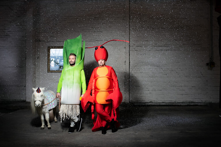 Ein Foto von Tilm Mälzer und Sasha, die in Hummer- und Lauchkostüm ein Zirkusponi an einer Leine halten.